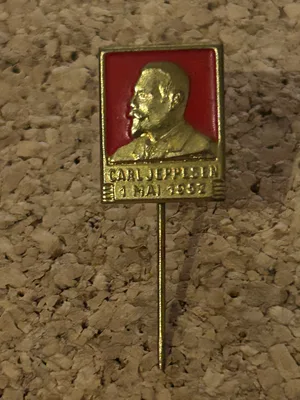 Det offisielle 1. mai-merket fra 1952 med et gullfarget relieffportrett av Carl Jeppesen mot rød bakgrunn.
