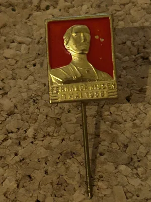 Rektangulært gullfarget 1. mai-merke fra 1956 med Fernanda Nissen i relieff mot rød bakgrunn.